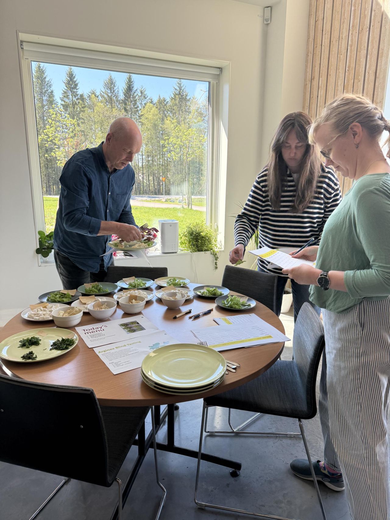 Three people are gathered around a round wooden table set with multiple plates and bowls containing various leafy greens and food samples. One man in a dark blue shirt is serving food onto a plate, while two women, one in a striped black and white sweater and the other in a green top with striped pants, are looking at papers, possibly tasting notes or a menu. The setting is a bright room with a large window showing trees and greenery outside. The table also has pens and printed sheets titled "Today's menu" and "Arevines Blind Tasting.