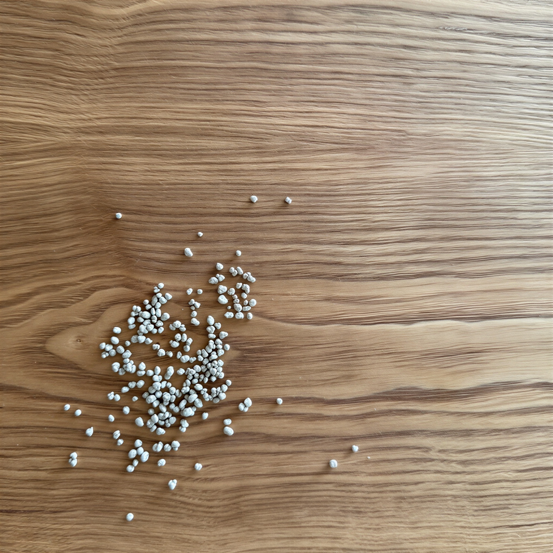 Small white granules scattered on a light brown wooden surface with a natural grain pattern.