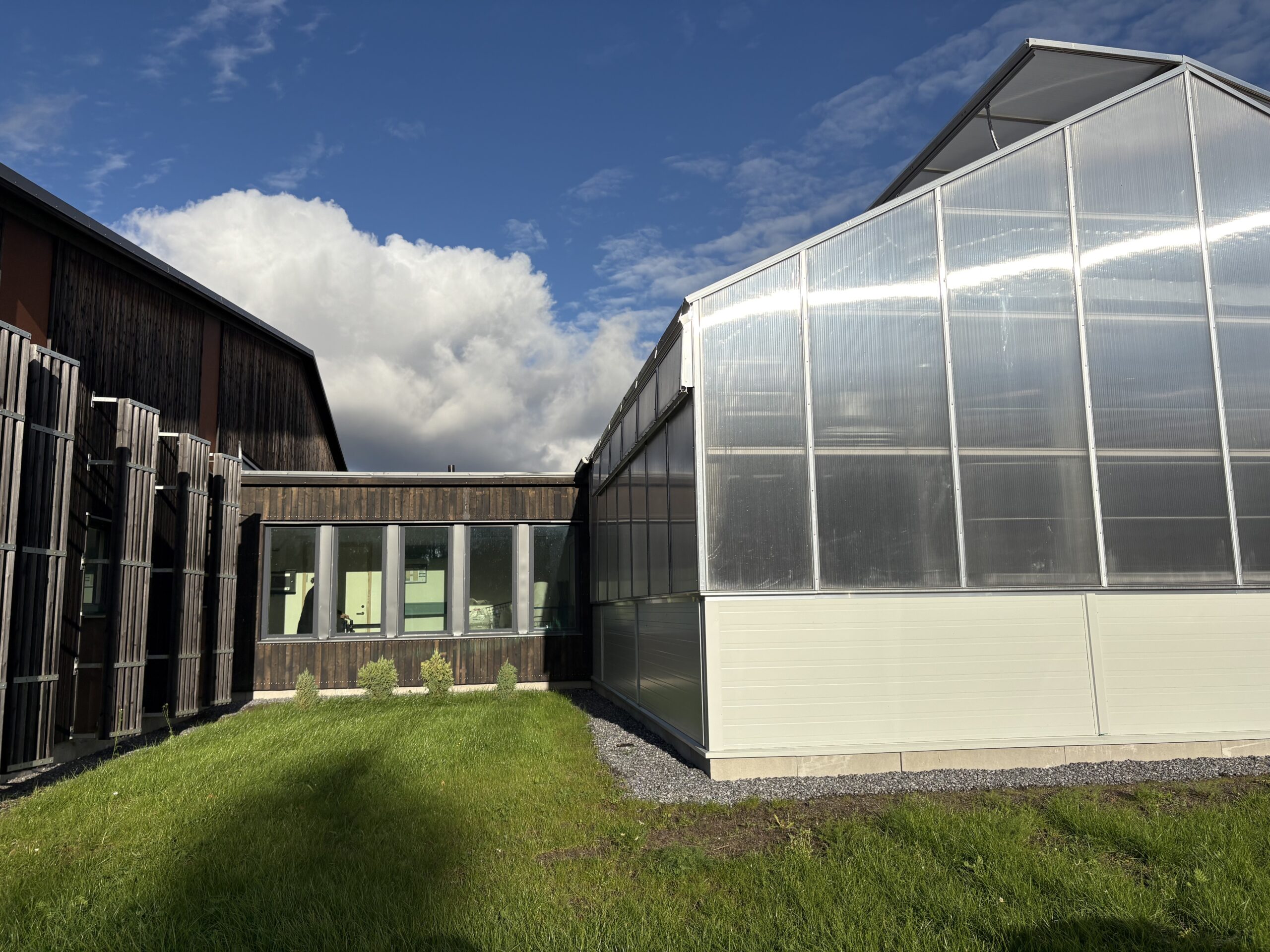 A modern building with dark wooden siding and large windows on the left, adjacent to a translucent greenhouse structure with a metal frame on the right. The foreground features a green grassy area with small shrubs, under a partly cloudy blue sky.