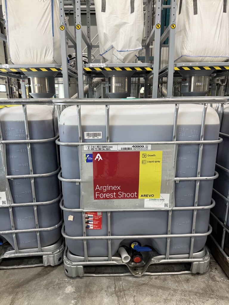 A large industrial container with a metal cage, labeled "Arginex Forest Shoot" by AREVO. The label indicates it is used for growth and liquid spray. The container is gray with measurement markings on the side and has a blue valve at the bottom. It is placed on a concrete floor in a warehouse setting, with other similar containers and large white bags visible in the background.